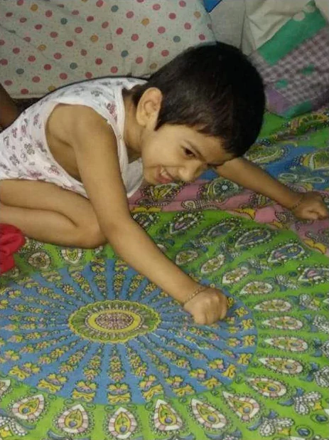 A smiling boy kneeling on a bright patterned bedsheet, tracing the pattern with his finger