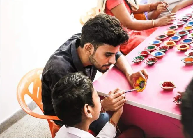 A young man and a boy painting clay diyas at a pink table