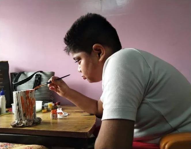 A teenage boy at a table, painting a craft piece with a fine brush