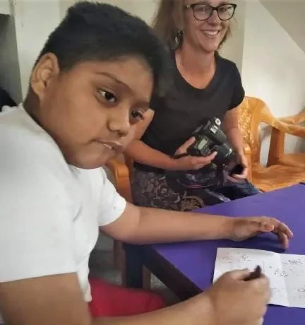 A boy writing at a purple table while a smiling volunteer holds a camera
